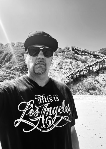 RAMIREZ, black and white portrait: cap and sunglasses, wearing a This is Los Angeles script tee, on a beach with a wooden staircase behind him